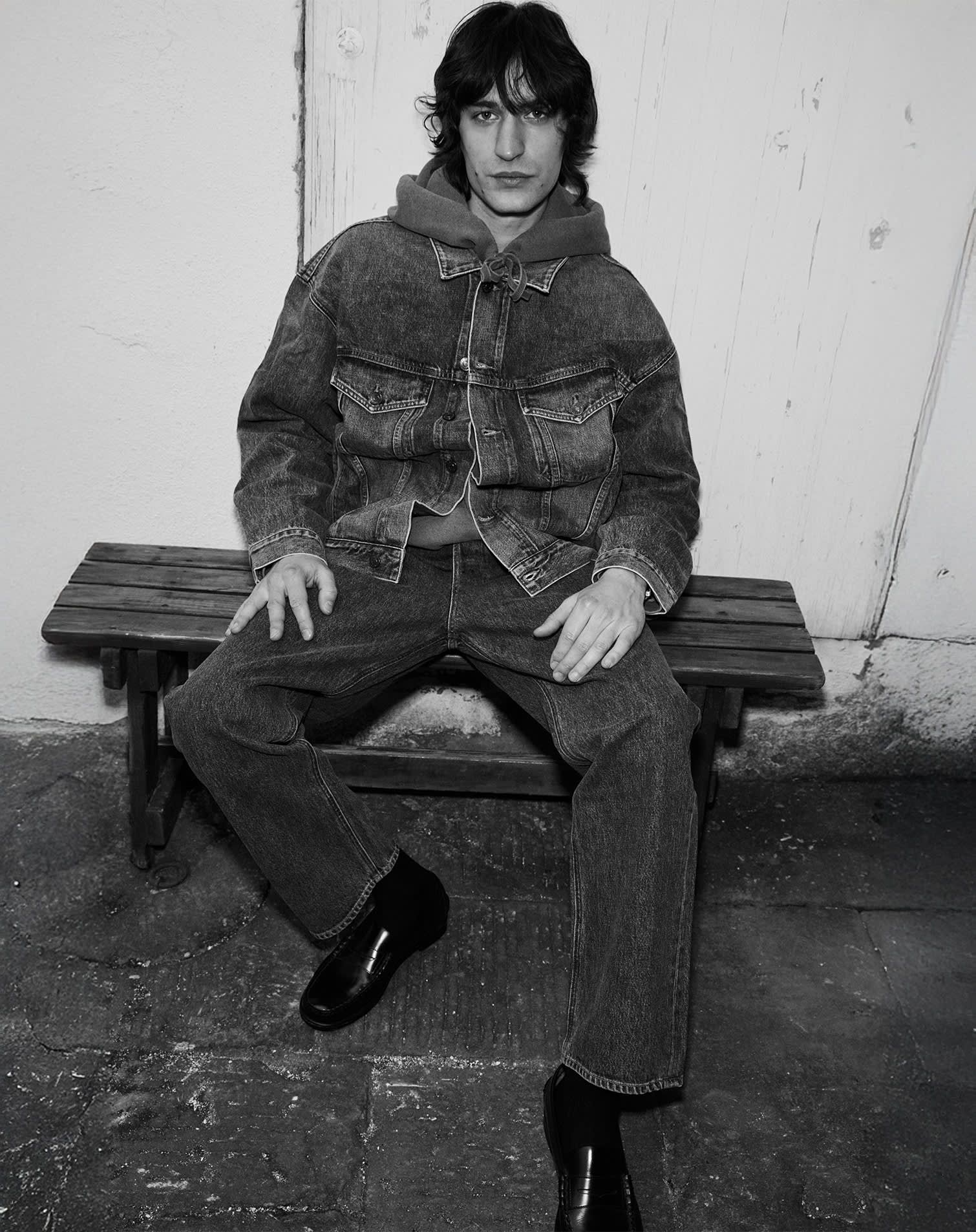 man wears jeans and denim jacket over hooded sweatshirt and black loafers