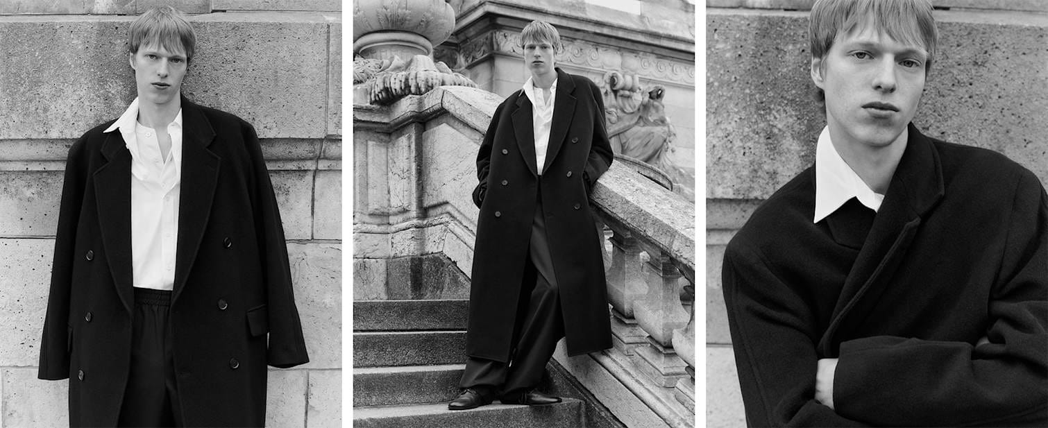 Black and white photo of man posing on stairs in Auralee wool coat, The Row wool jogger pants and Marsell shoes.