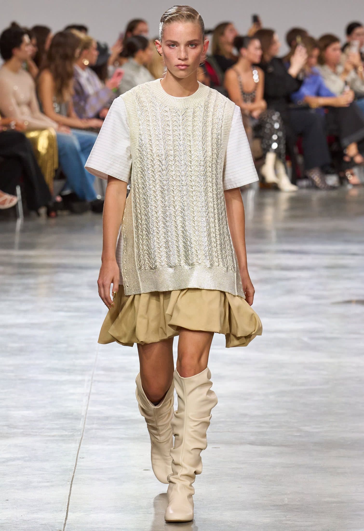 runway model with knit sweater vest over white t-shirt, beige knee-high boots and dark beige skirt